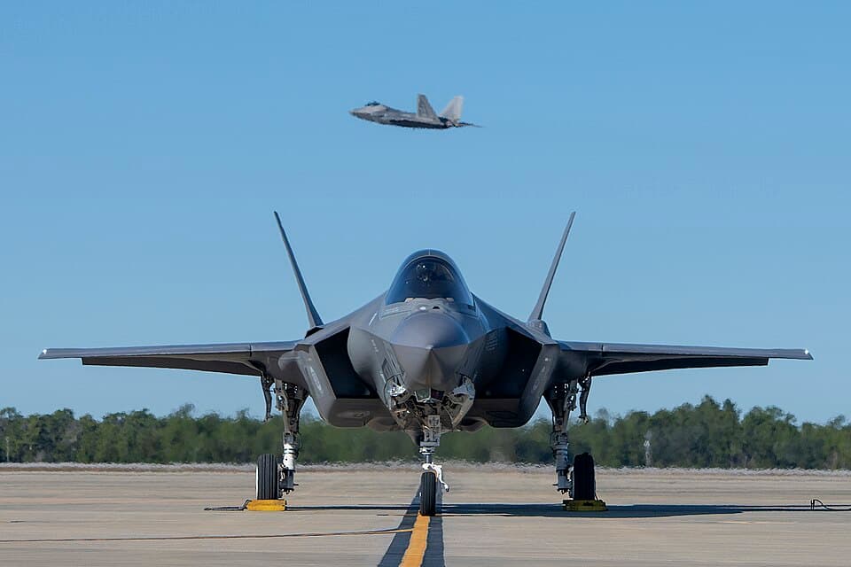 Front view of an F-35A on a taxiway, with an F-22 flying in the background